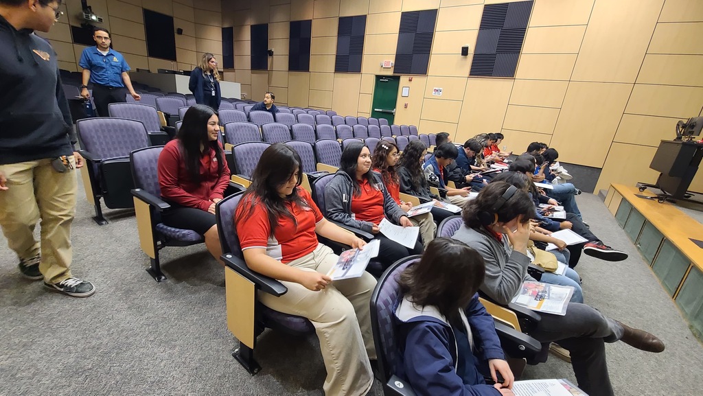 A group of students sits in chairs in a lecture hall, holding papers and listening to a person standing.