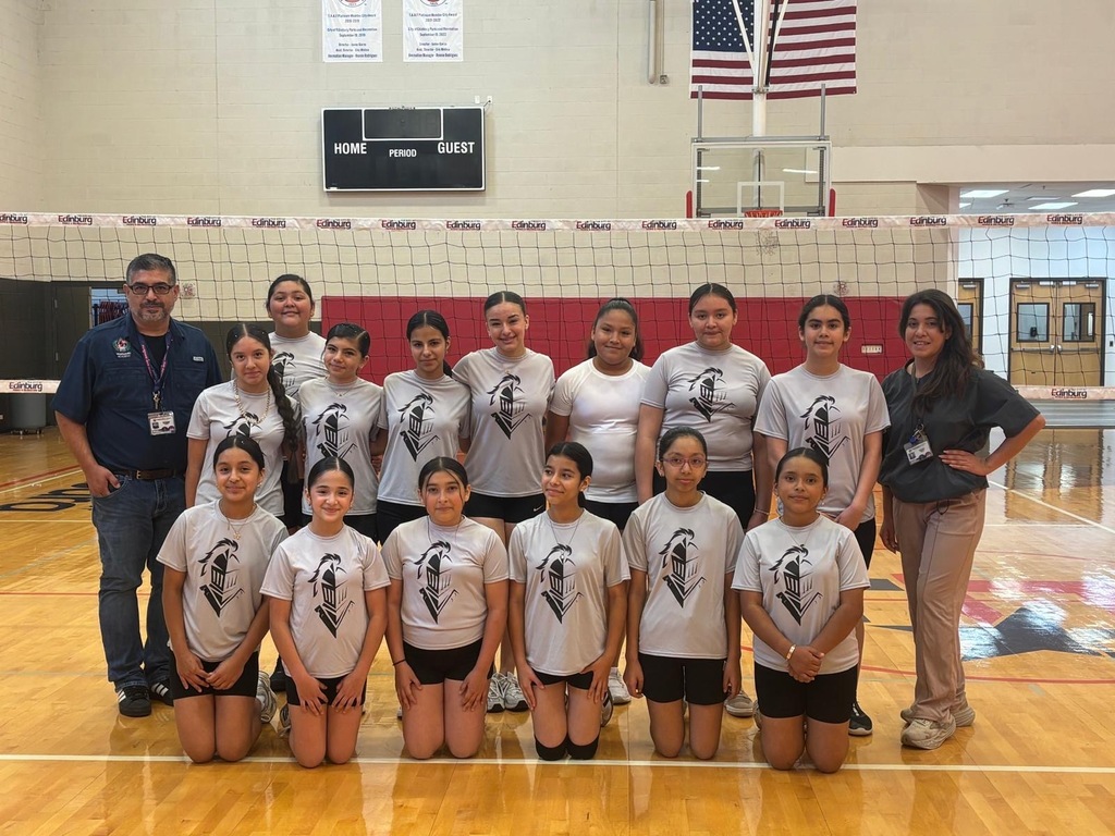 A volleyball team poses for a group photo in a gym. They wear matching shirts and shorts. A coach stands behind them.