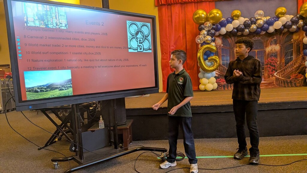 Two children standing in front of a large screen displaying an image, with one speaking and the other listening.