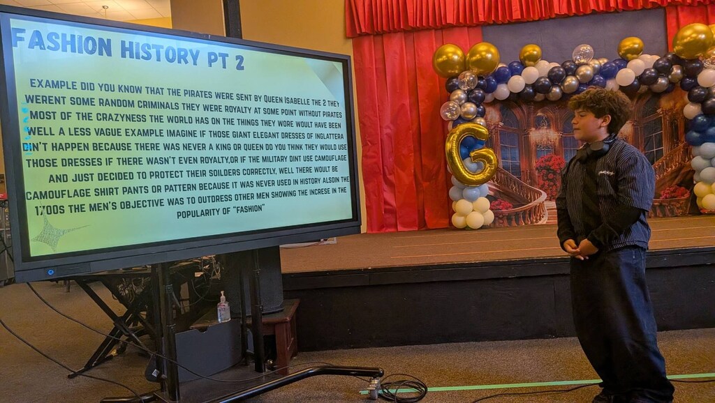 A person stands before a monitor with "Fashion History Pt 2" on a stage with balloons.