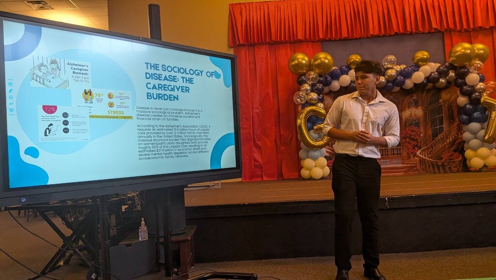 A person stands on stage with a large screen displaying text about a disease. Behind, balloons are tied to a red curtain.