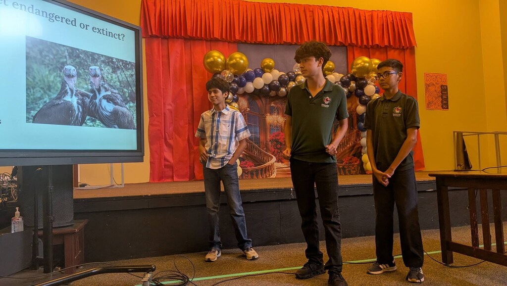 Three boys on a stage with a projector screen behind them showing an image. The stage is decorated with red fabric.