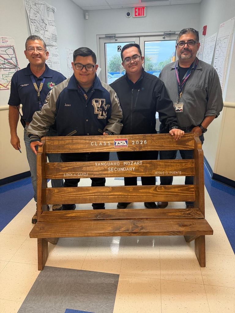 Four men pose behind a wooden bench with engraved text in a hallway. The men wear glasses, jackets, and lanyards.