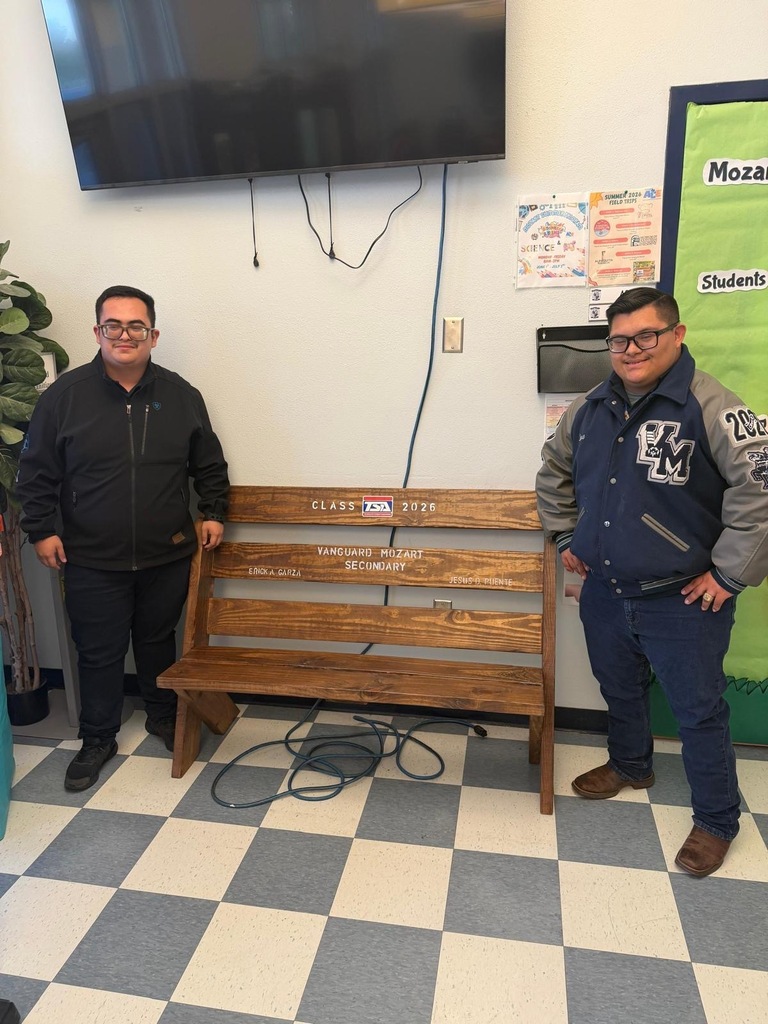 Two men in jackets stand beside a wooden bench with engravings, against a backdrop of a TV, bulletin board, and checkered floor.