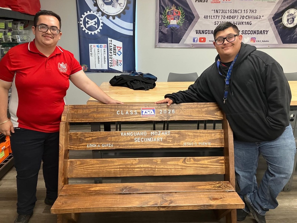 Two men stand behind a wooden bench, one in a red shirt and the other in a black jacket.
