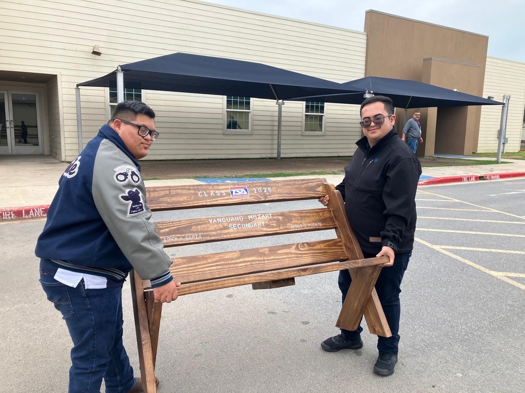 Two men, one in glasses, lift a wooden bench in a parking lot with a building and canopy tents in the background.
