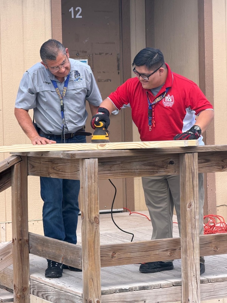 Two men in a workshop. One holds a yellow sander and the other wears a red shirt.