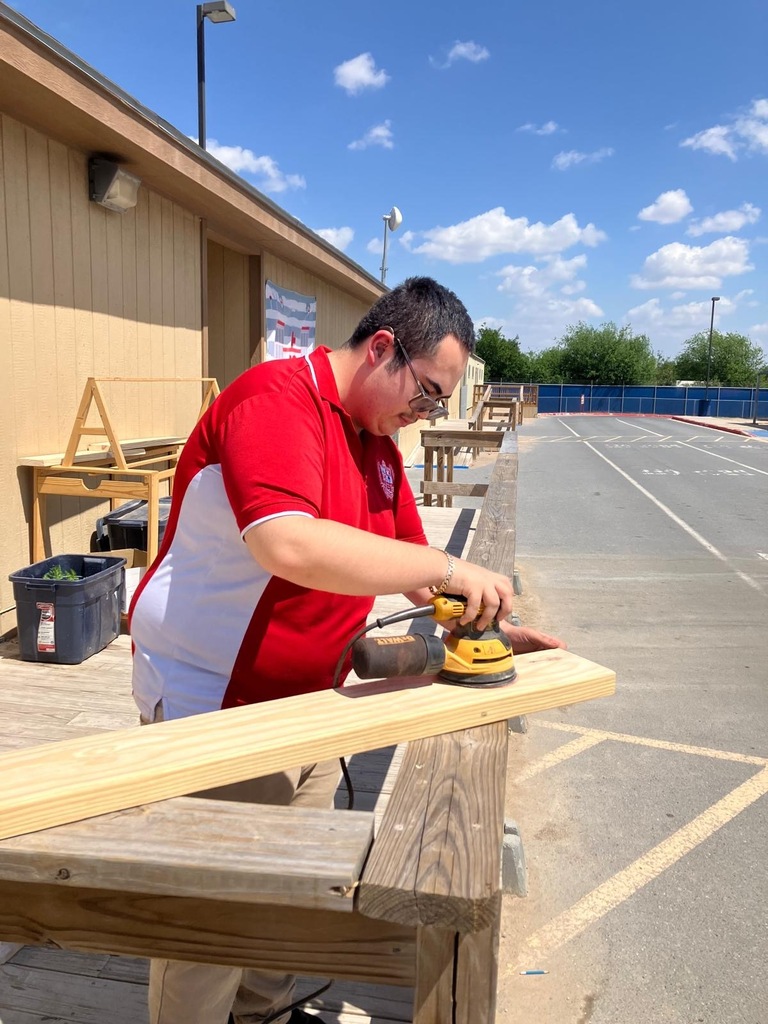 A person uses an electric sander on a wooden plank. A building and parking lot are in the background.