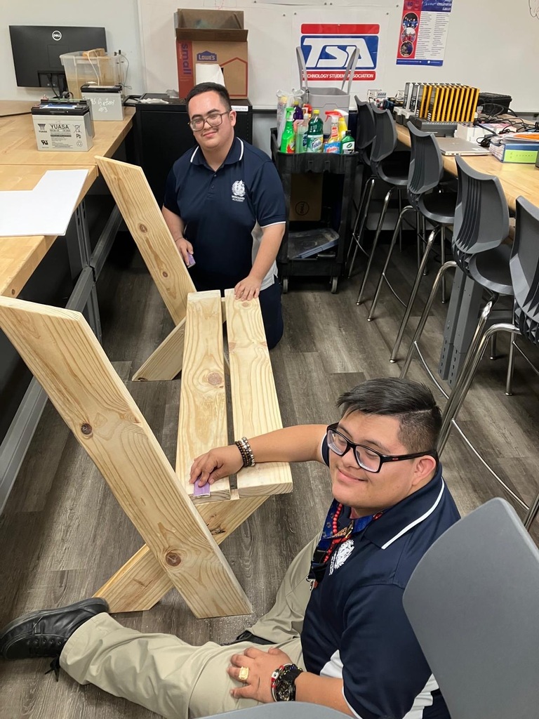 Two people wearing uniforms and glasses work on a wooden bench in a room with tables.