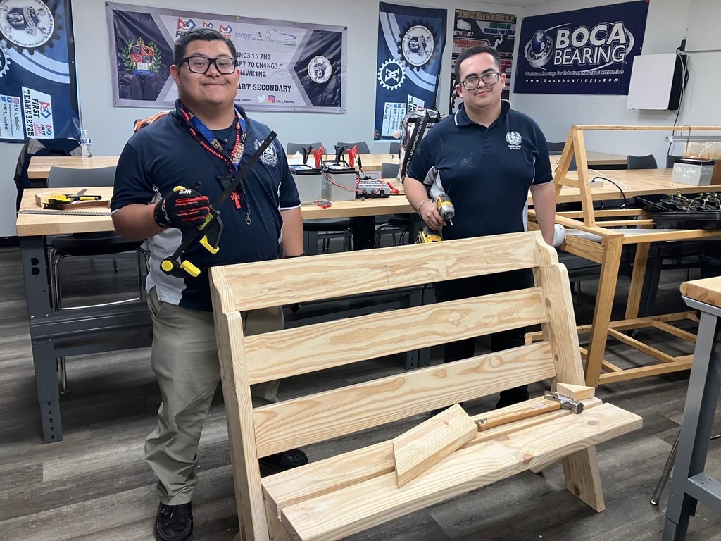 Two men in blue shirts and glasses stand next to a wooden bench in a workshop. Behind them, banners and tables are visible.
