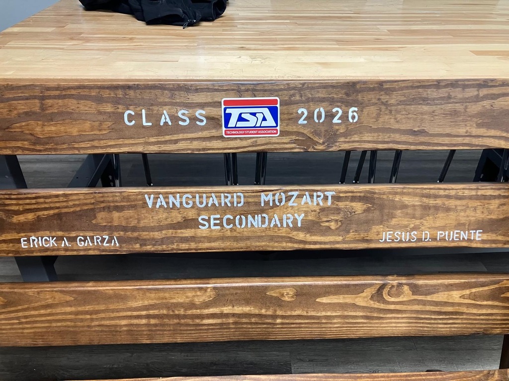 Wooden table with engravings. Top has "CLASS TSA 2026" and a logo. Two benches below, one with "Vanguard Mozart Secondary" and "ENRICA GARZA," the other with "JESUS O PUENTE."
