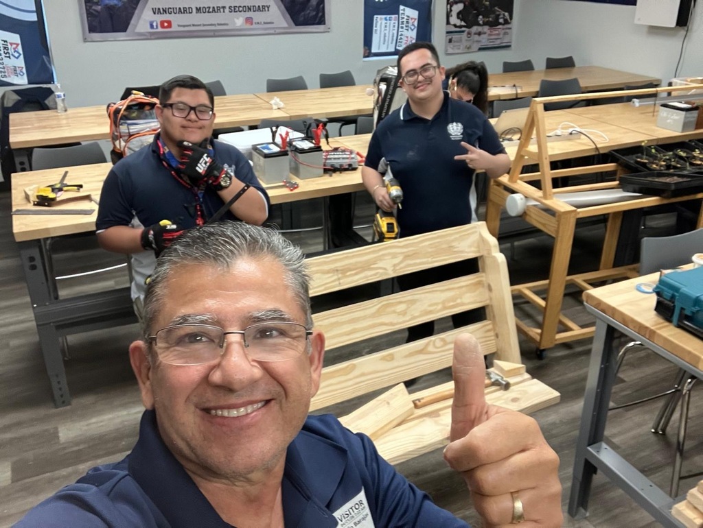 Four men in a workshop, one smiling and giving a thumbs up. Wooden tables and benches. A poster in the background.