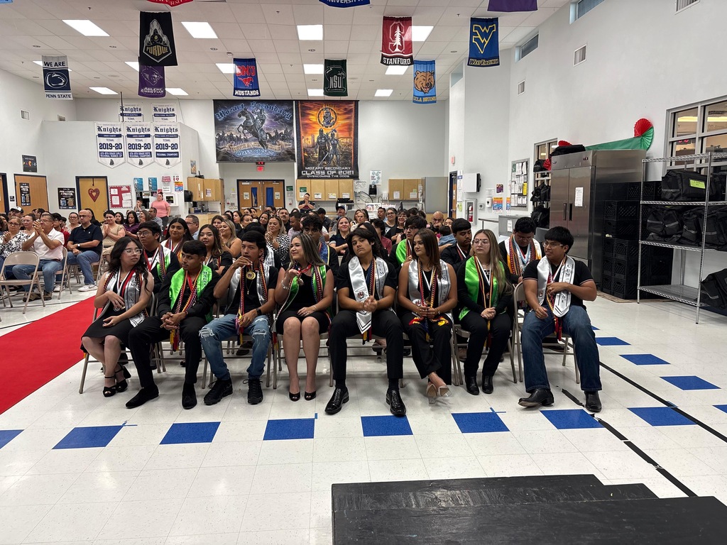 A group of students wearing green sashes sit on chairs in a room with white walls.