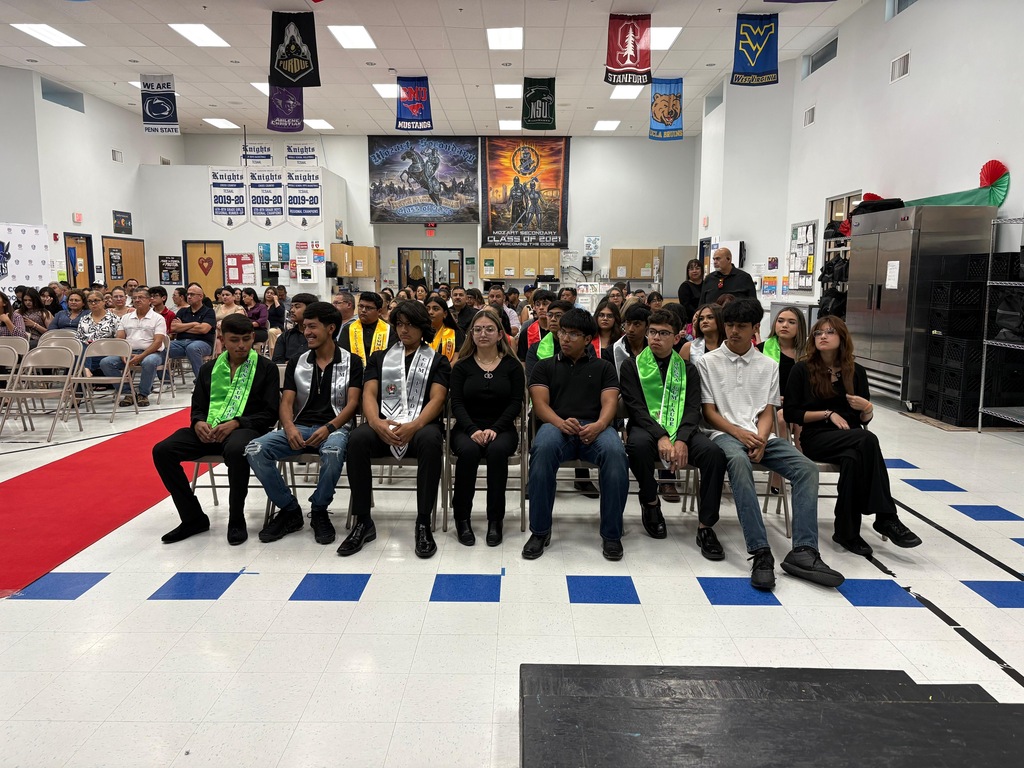 A group of people wearing green sashes sit on chairs in a room with blue and white squares.