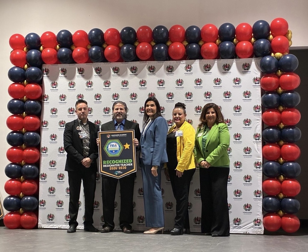 Five people stand on a stage with a backdrop of blue and red balloons, one holds a plaque.