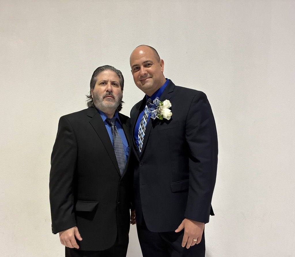 Two men in suits and ties stand next to each other against a plain white background.
