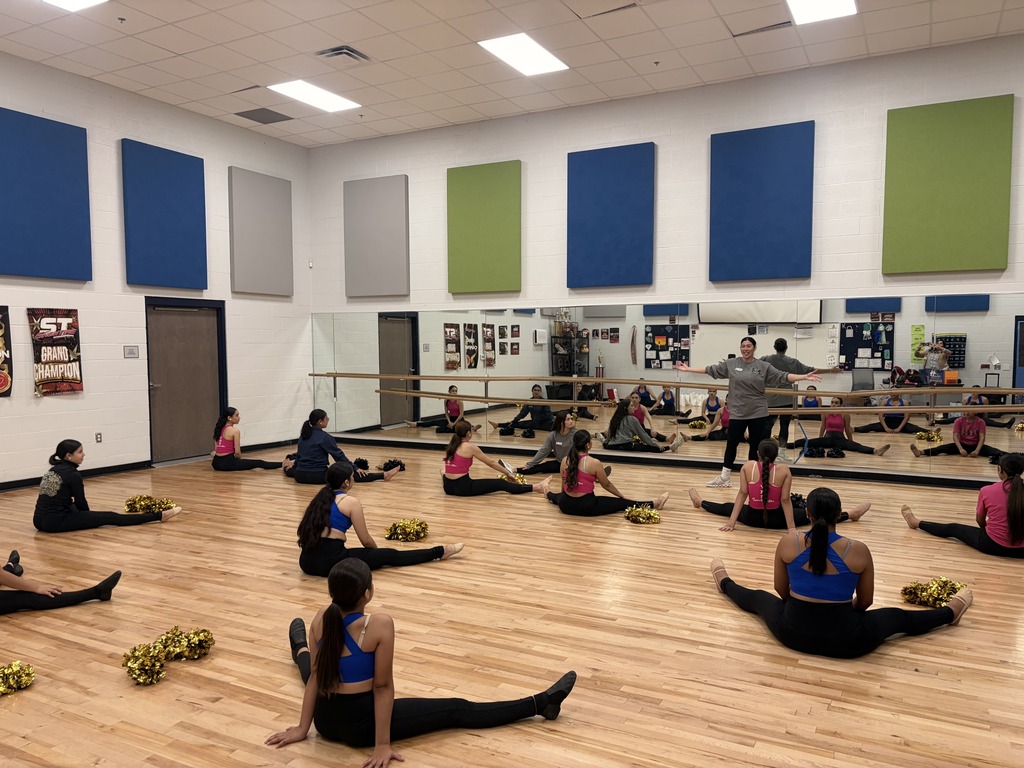 A dance studio with wooden floors and blue walls. Several women are seated on the floor, engaged in stretching exercises.