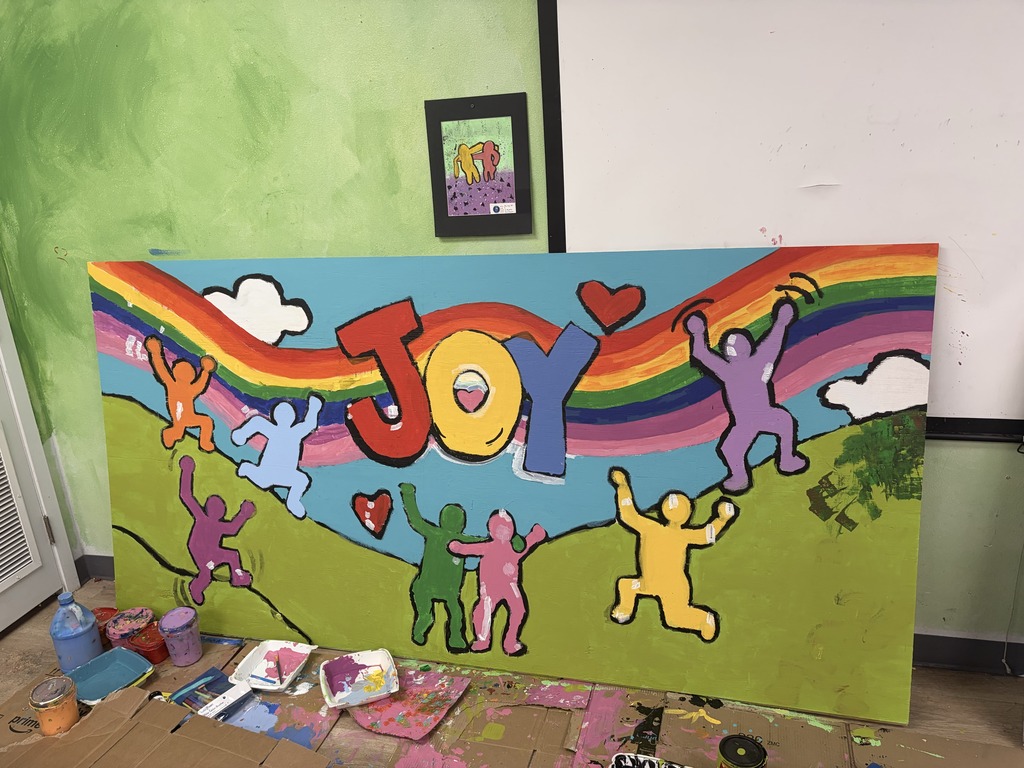 Large painting of smiling figures and a rainbow with the word "JOY" in front of green wall.