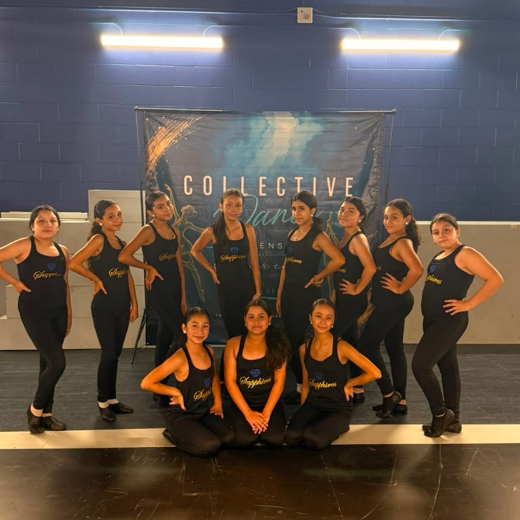 A group of dancers in black attire pose for a photo in a studio with a banner.