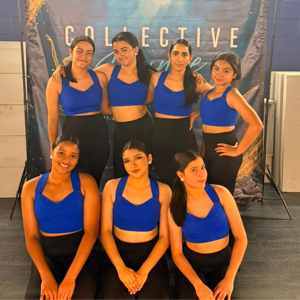 Six women in blue tops and black pants pose for a photo in a dance studio.