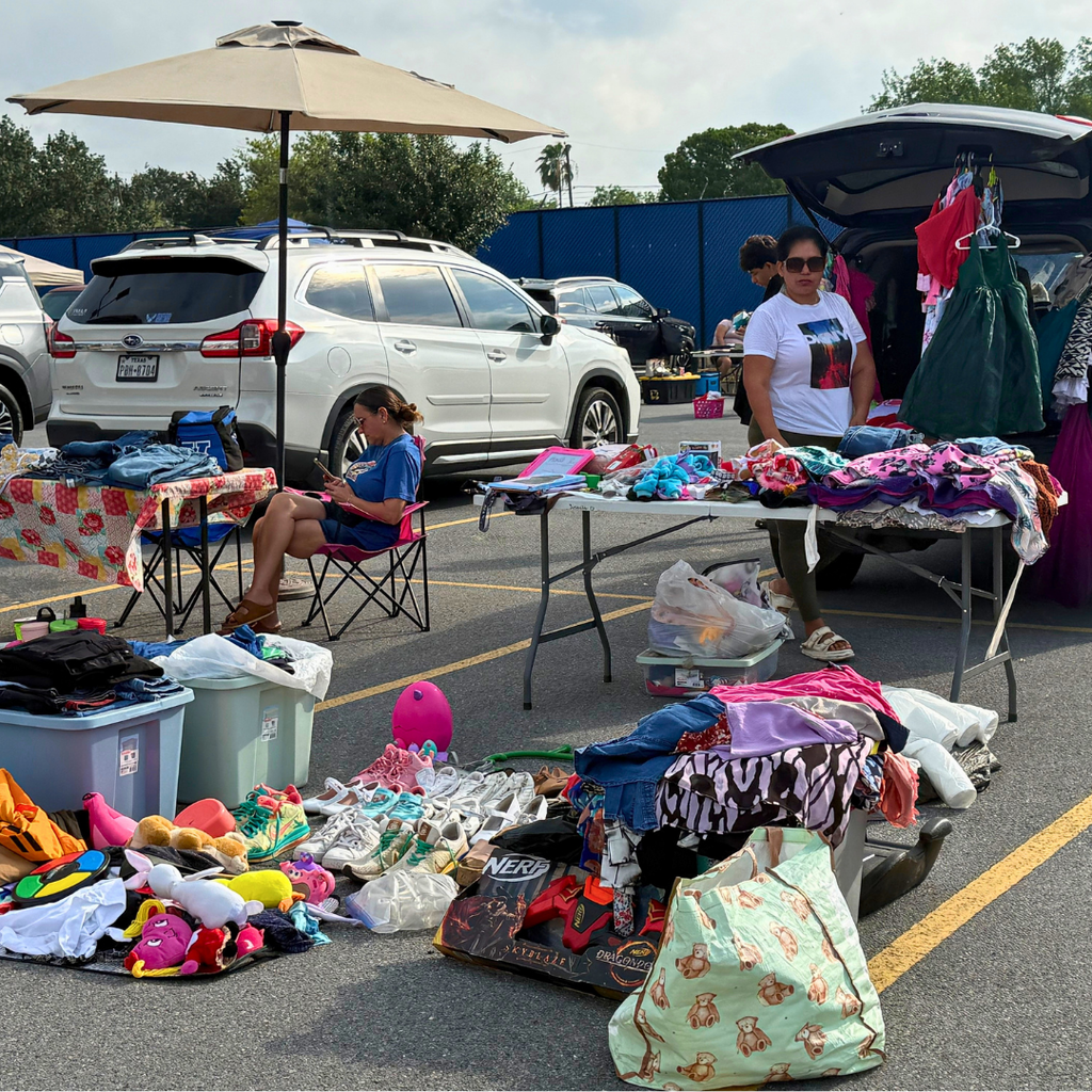 Tables with clothing, toys, and accessories are set up in a parking lot. A person sits in a chair.