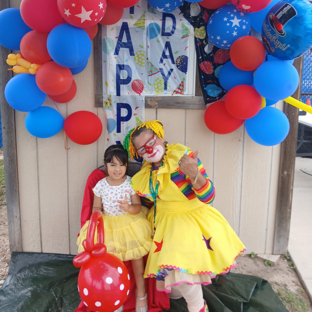 A clown and a child pose for a photo in front of a wall with balloons.