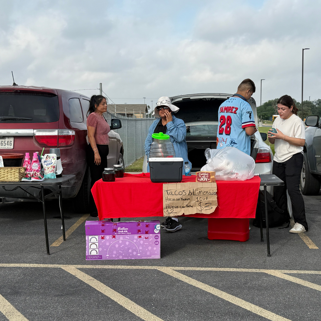 Four people near a table with food items on it, with cars parked behind them in a parking lot.