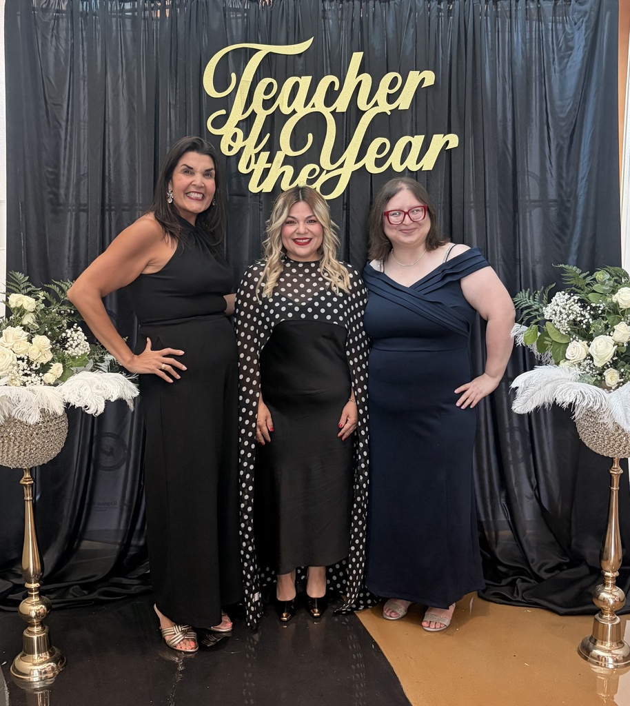Three women in evening dresses stand in front of a black backdrop with a gold banner. White flowers are in vases.