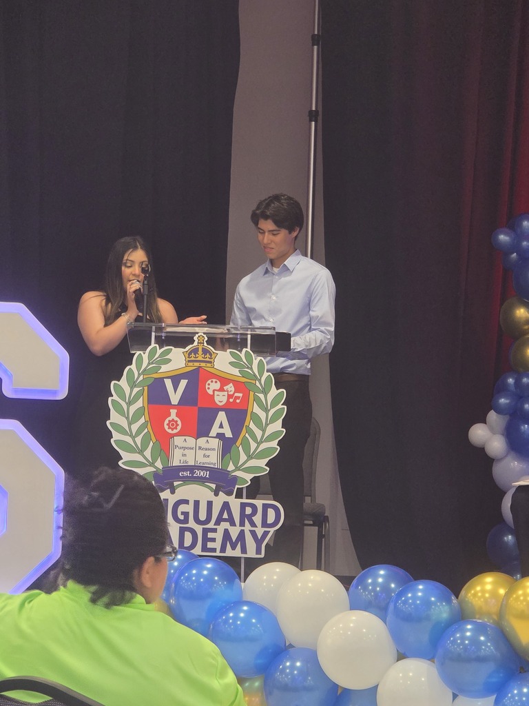 A person speaks at a podium. A large emblem sits behind them. Behind and to the right, balloons in blue and white.
