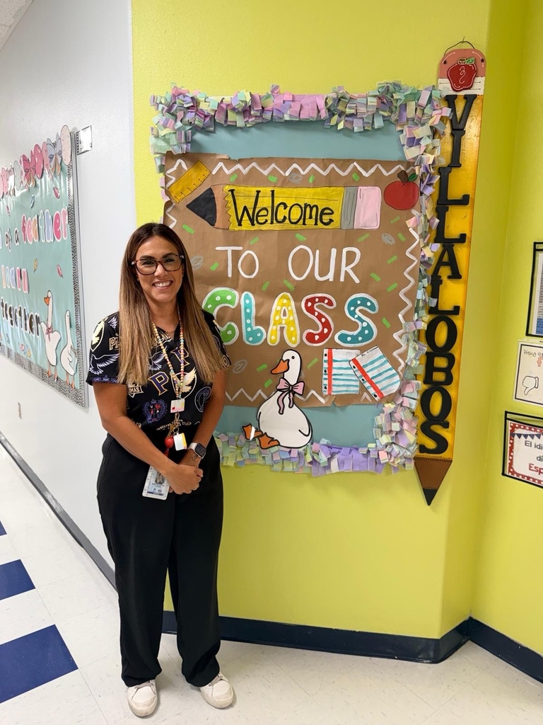 A woman stands next to a yellow classroom wall with a "Welcome to Our Class" sign.