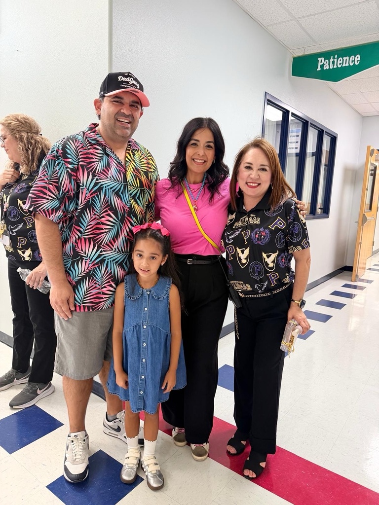 Four people stand together in a hallway, smiling. The man wears a colorful shirt and cap, and the girl wears a blue dress. The two women wear pink and black tops.