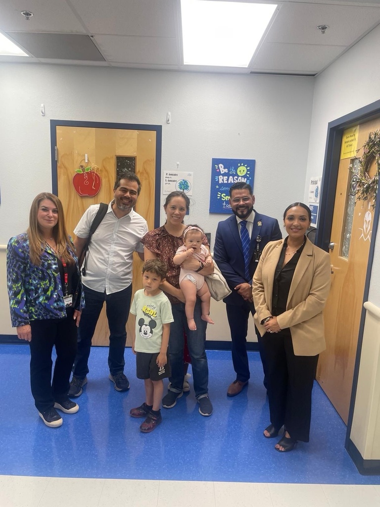 A group of people, including adults and children, are standing in a hallway. A baby is held by an adult.