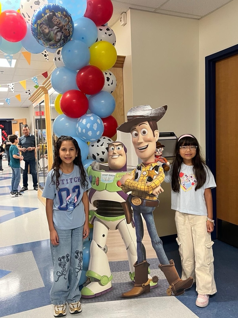 Two girls stand next to Woody and Buzz Lightyear character mascots in a corridor with colorful balloons.