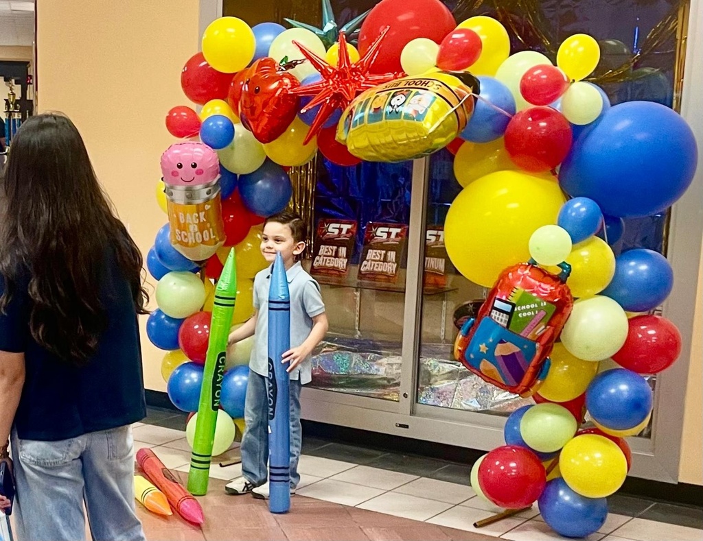 A child stands in front of a colorful balloon arch, holding a crayon and balloon. A woman with long hair stands nearby.