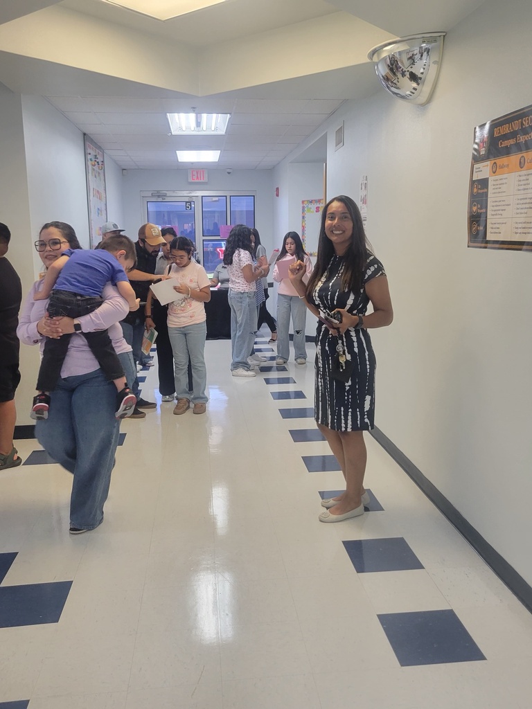 A hallway with blue squares on the floor. Several people are standing or walking. A woman holds a child on her shoulder.