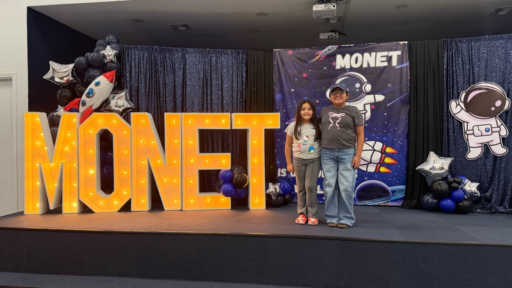 Two people on a stage with a large sign reading "MONET" in yellow lights. Background features blue curtains and balloons.