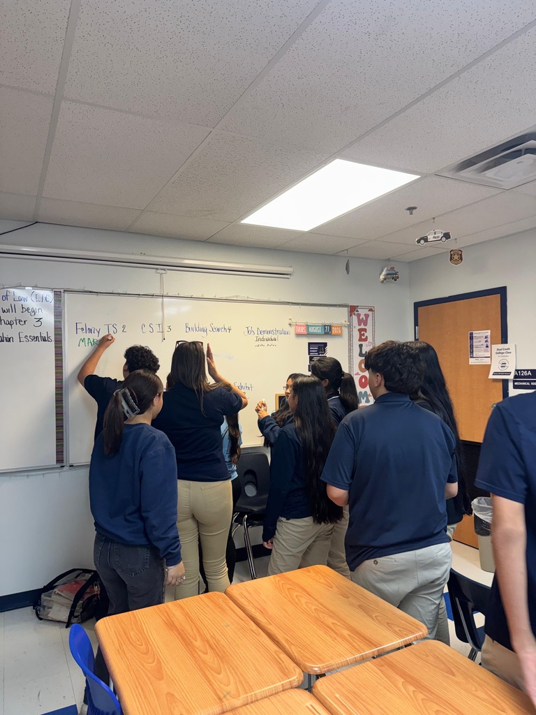 Group of students, wearing blue shirts, gathered around a whiteboard in a classroom. Some are writing on it.