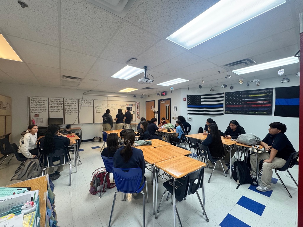 A classroom with students sitting at desks. The room has white walls, blue floor markings, and ceiling lights.
