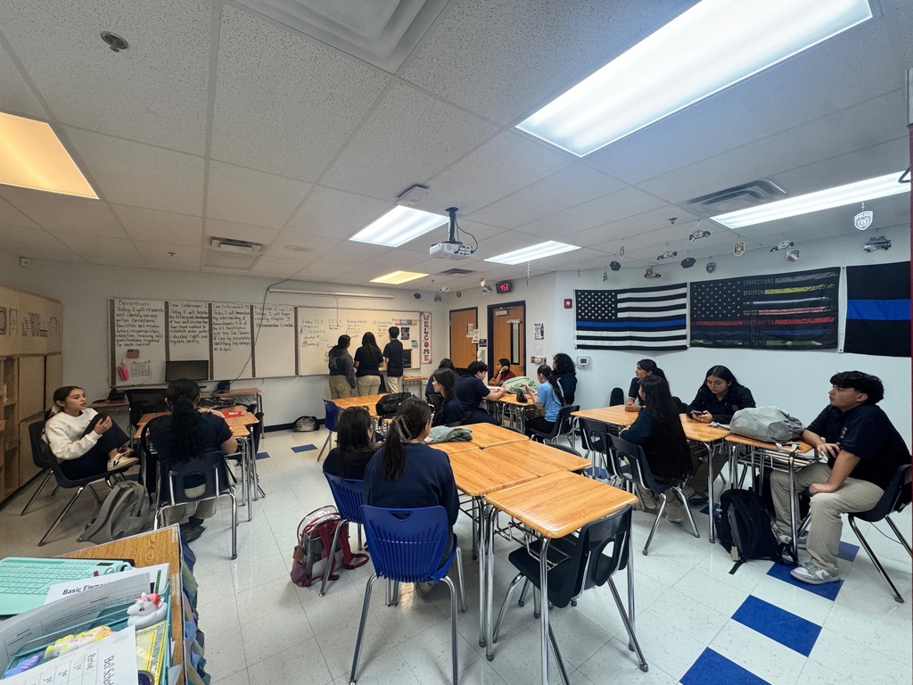 A classroom with students seated at tables, with an American flag on the wall, and overhead lighting.