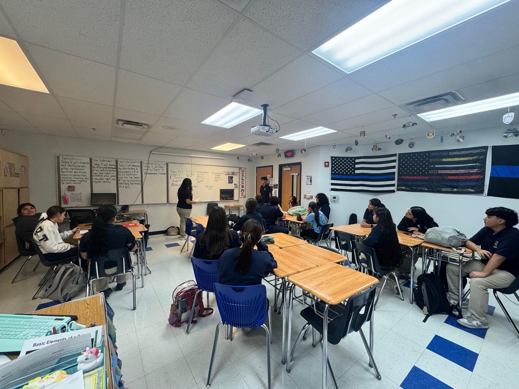 Classroom with desks, chairs, a teacher, and flags on the wall. Students seated, working or listening. White walls, ceiling lights, and blue floor stripes.