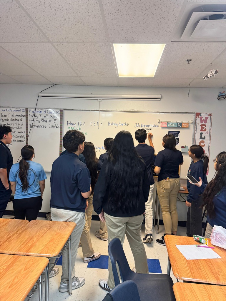 A classroom with people standing around a whiteboard; others stand near desks. A light fixture is on the ceiling.