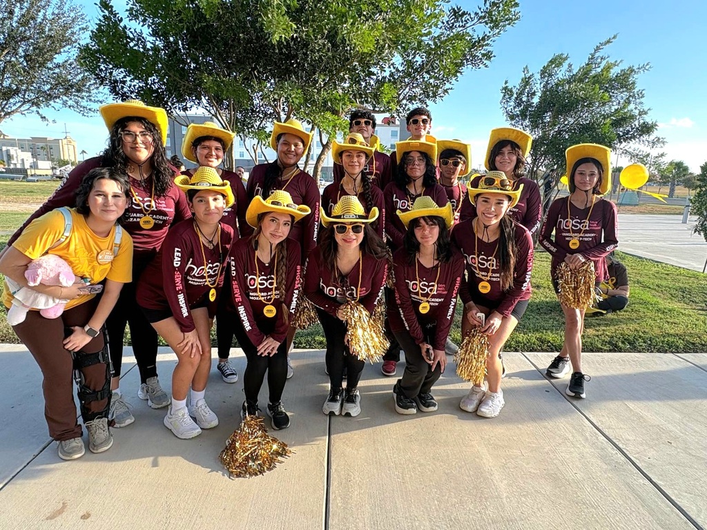 A cheerleading team dressed in maroon uniforms, yellow cowboy hats, and white sneakers stands on a sidewalk with trees in the background.