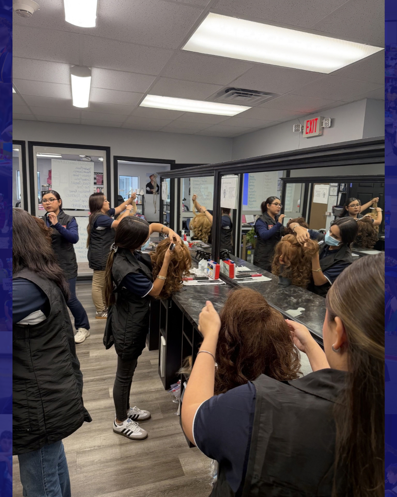 Multiple women in a salon, wearing black vests, standing and working on hair. Some are using hair dryers.