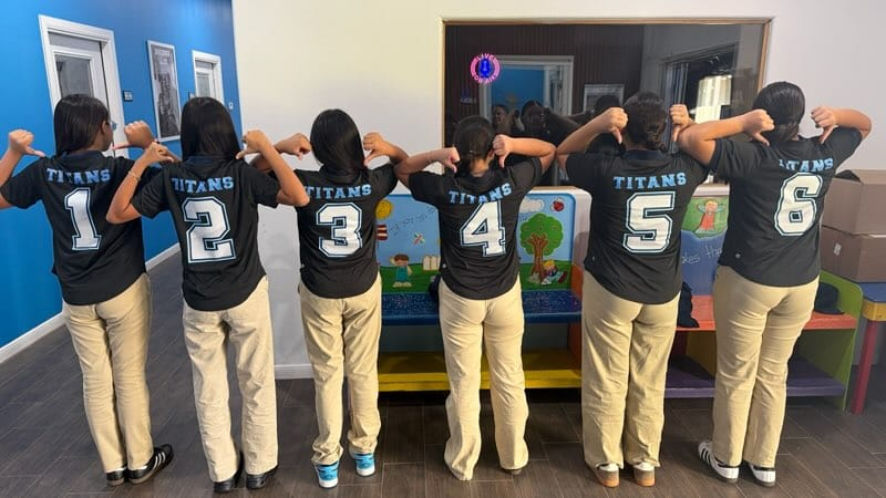A group of women in numbered black shirts pose in a room with blue walls, white frames, and a colorful bench.