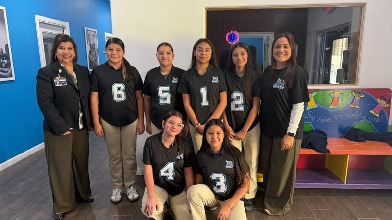 A group of women in numbered black shirts pose in a room with blue walls, white frames, and a colorful bench.