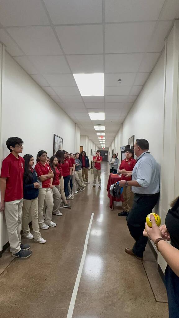 A hallway with white walls and ceiling. People in red shirts stand along one wall. A man in a light blue shirt speaks to them.