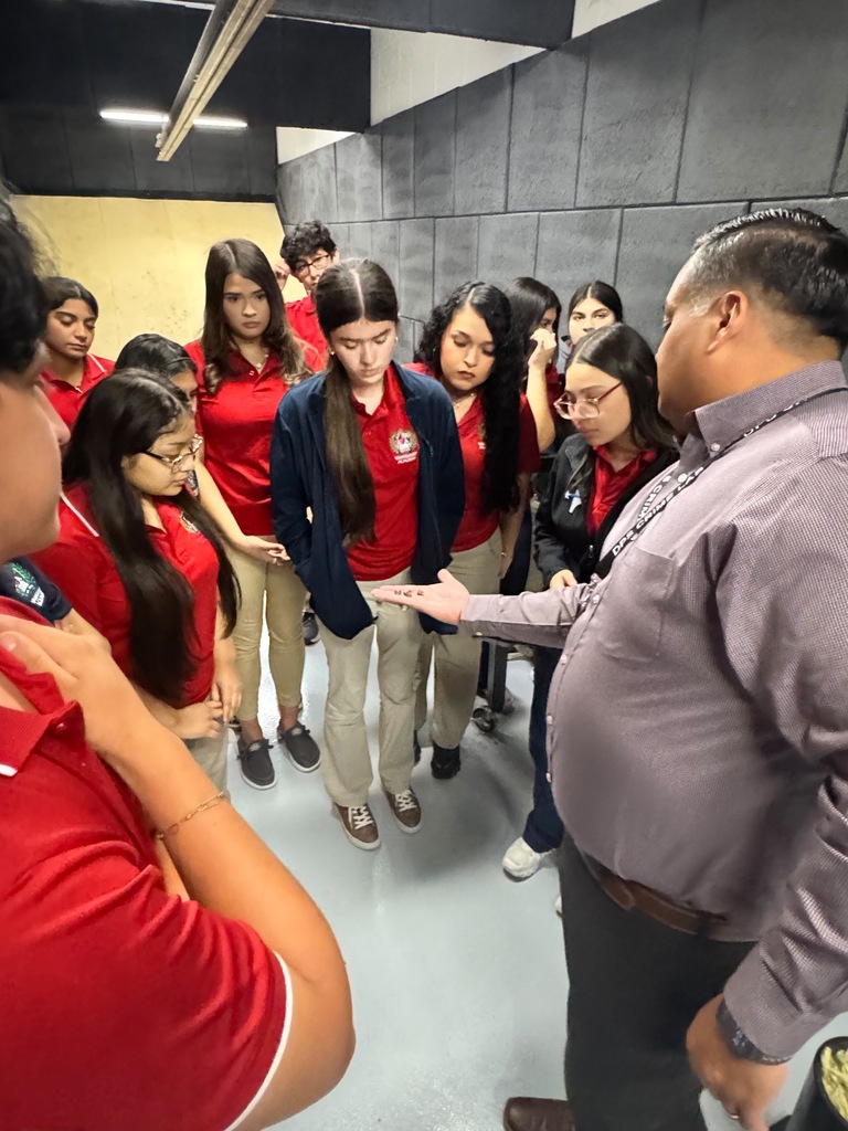 Students in red shirts and khaki pants gather around a man in a gray shirt. They are indoors.
