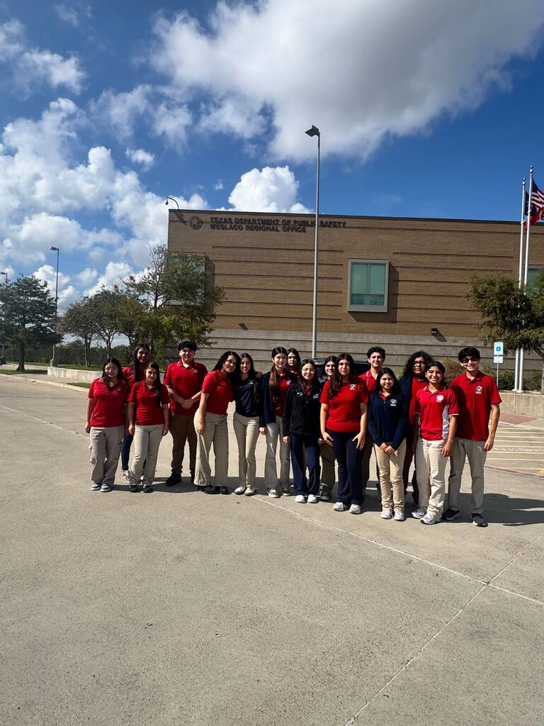 A group of students in matching red shirts and pants stand in a parking lot in front of a building.