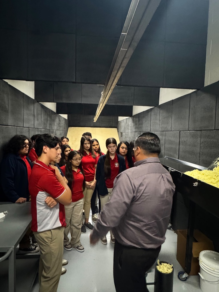 A group of students wearing red shirts and pants stand in a room with a man explaining something.