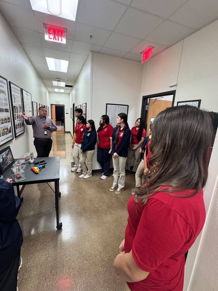 A group of individuals dressed in red shirts and khaki pants stand in a hallway. A person speaks to the group.